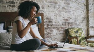 Woman drinking coffee while using laptop in bed.
